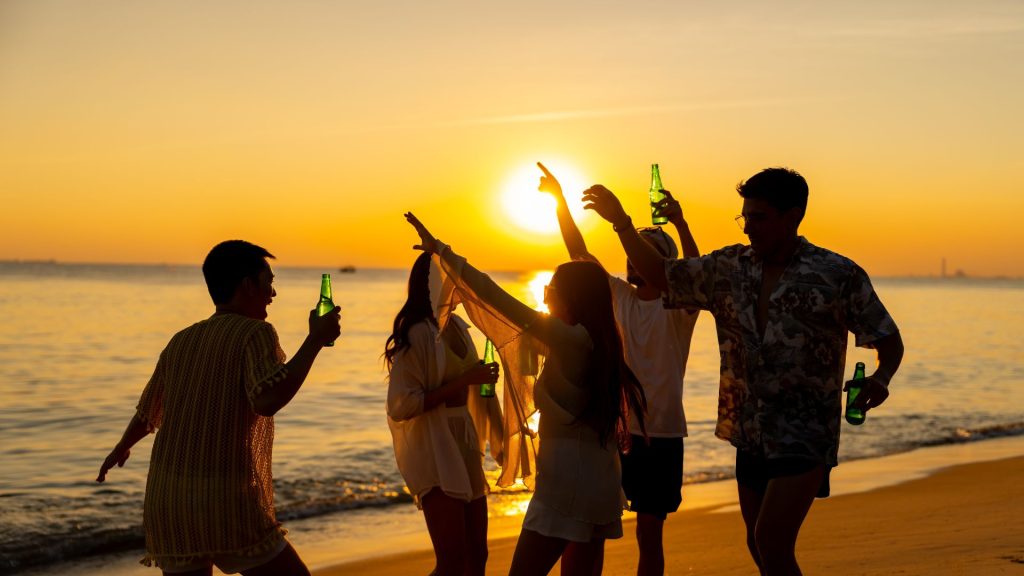 Image of people dancing on a beach at sunset, holding green bottles, with the sun low over the ocean.